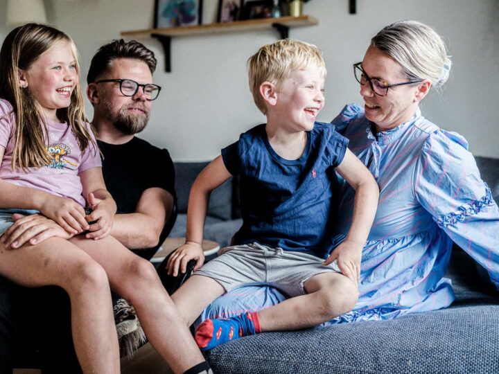 Billedet viser en familie bestående af to voksne og to børn. De to børn sidder på skødet af deres far og mor. Hele familien griner